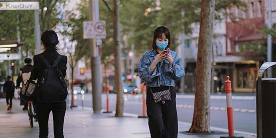  A young women walking on a sidewalk in an Australian city during the day. She is wearing a blue surgical mask and is looking down at her phone. The surrounding street looks empty with a few people wandering, it feels reminiscent of the empty CBD in lockdown times.  