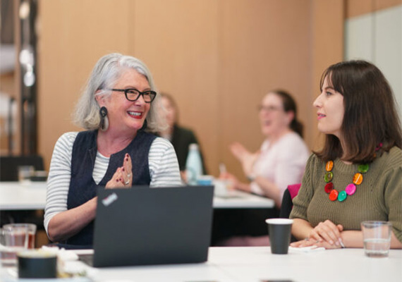Two women speak with a laptop open on the table.