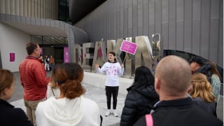 A student stands in front of a small crowd holding a pink sign for a campus tour at Deakin Open Day.