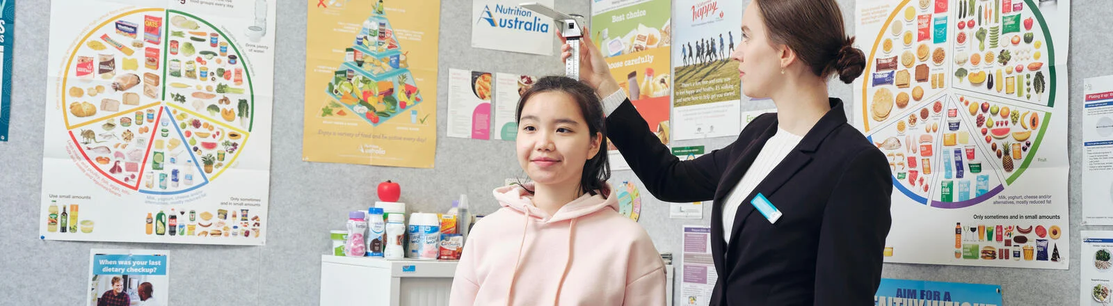 Deakin master of dietetics student measures height of patient in medical office with nutrition posters.