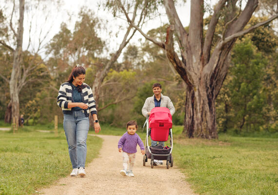 A mother walks in the park with a toddler. The father pushes a pram behind them.