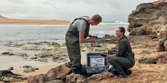 Two Marine Biology students crouch at the shoreline with equipment. They are wearing overalls.