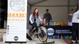 A person rides a stationary bike outside a stall for a DUSA club at Deakin Open Day.