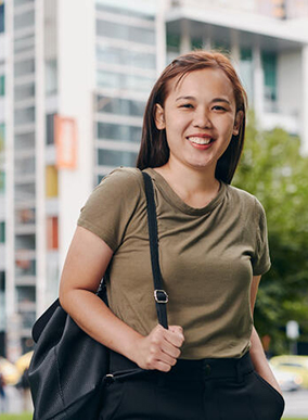 A smiling Deakin graduate students stands holding their bag outside Deakin Melbourne Burwood Campus.