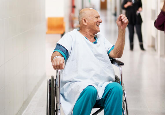 An ederly person in a hospital gown smiling and travelling in a wheelchair.