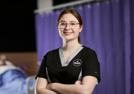 Nursing student Beth Lightbody looks at the camera with their arms crossed. They are wearing a nursing uniform.