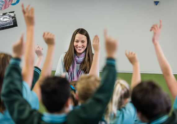 Primary school classroom, students with their hands raised, teacher at front.