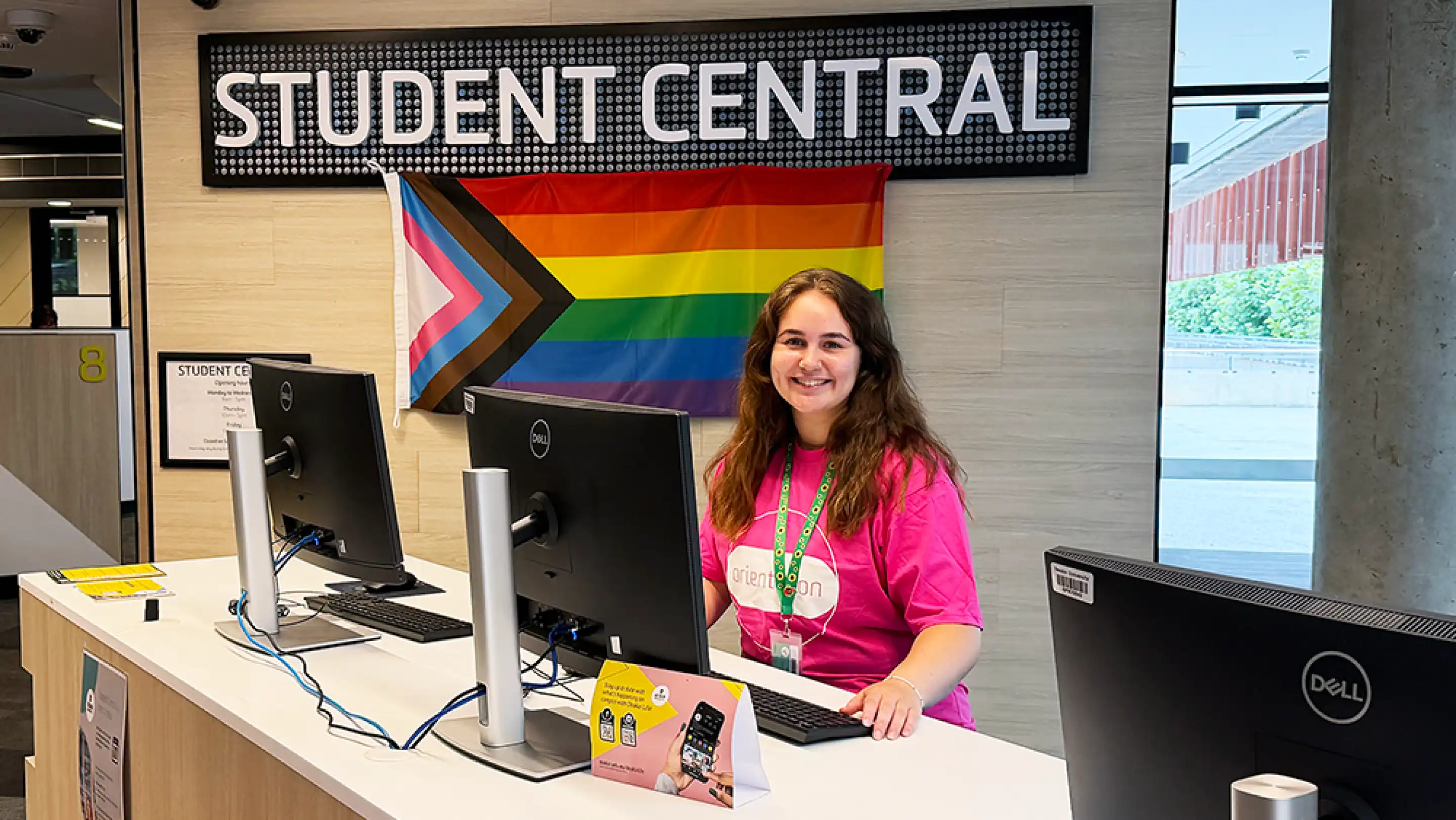 Image of a friendly staff member at the Student Central counter wearing a sunflower lanyard and in front of the pride flag