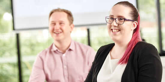 Two people holding pens and smiling inside a classroom.