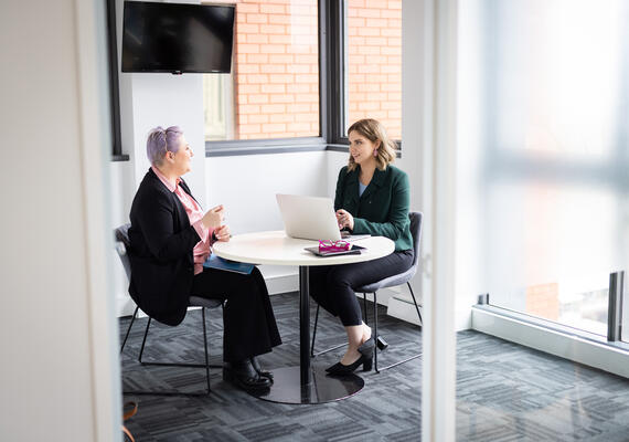 Two people sit across from one another at a desk and talk.