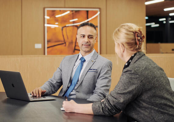 Two business people sit at a table with an open laptop in discussion.