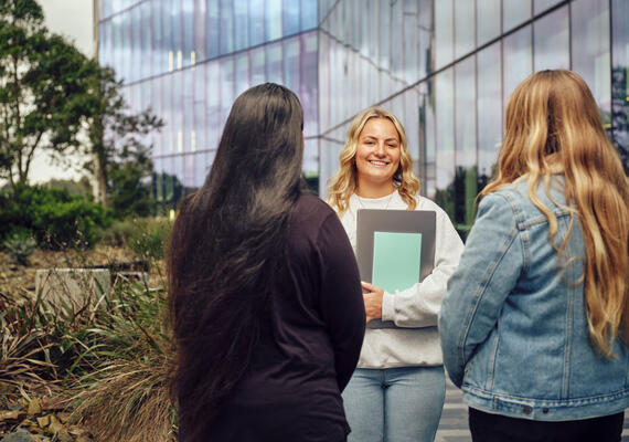 Smiling student holding books and standing in front of a large glass building.
