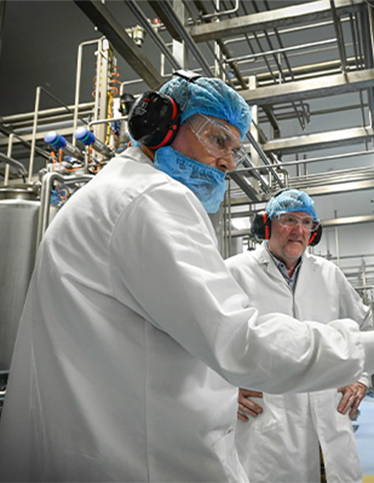 Two people working in a food processing facility, wearing lab coats, masks and ear protection.
