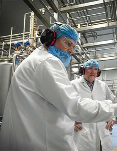 Two people working in a food processing facility, wearing lab coats, masks and ear protection.