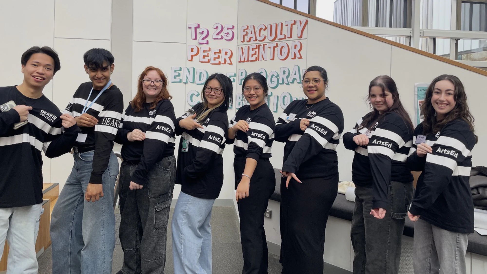 Gab and seven other peer mentors posing in a line, pointing to the Deakin logo and the word ArtsEd on their sleeve