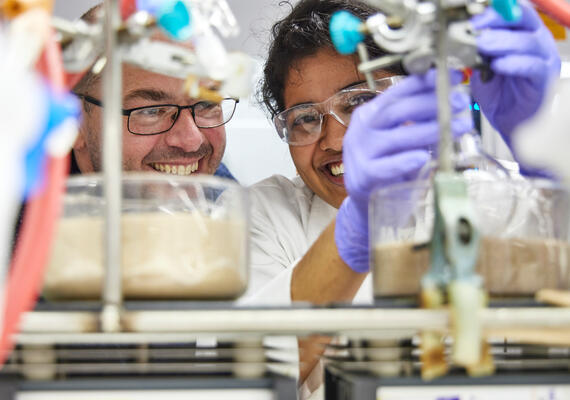Two research students in a lab, hold equipment while smiling to camera.