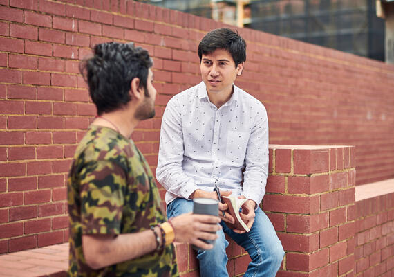 Two male adults sitting on a brick wall talking to each other