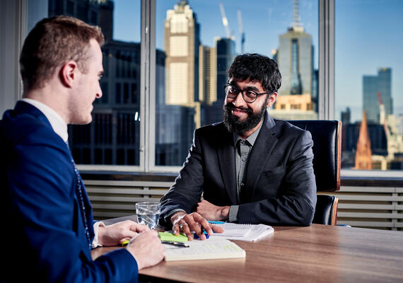 Two students in business attire, sitting either side of a desk in an office with a city view.