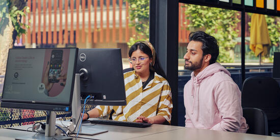 Two Deakin students sitting at a computer in a bright, modern workspace with large windows and leafy views, working together on a digital project.