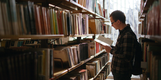 Deakin philosophy student standing in aisle of library, reading a book.