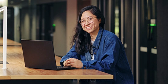 A Deakin student smiling while working on a laptop at a long wooden desk in a modern, well-lit study space.