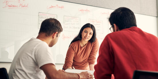 Three Deakin students in a meeting room, surrounded by papers, with notes written on the whiteboard behind them.