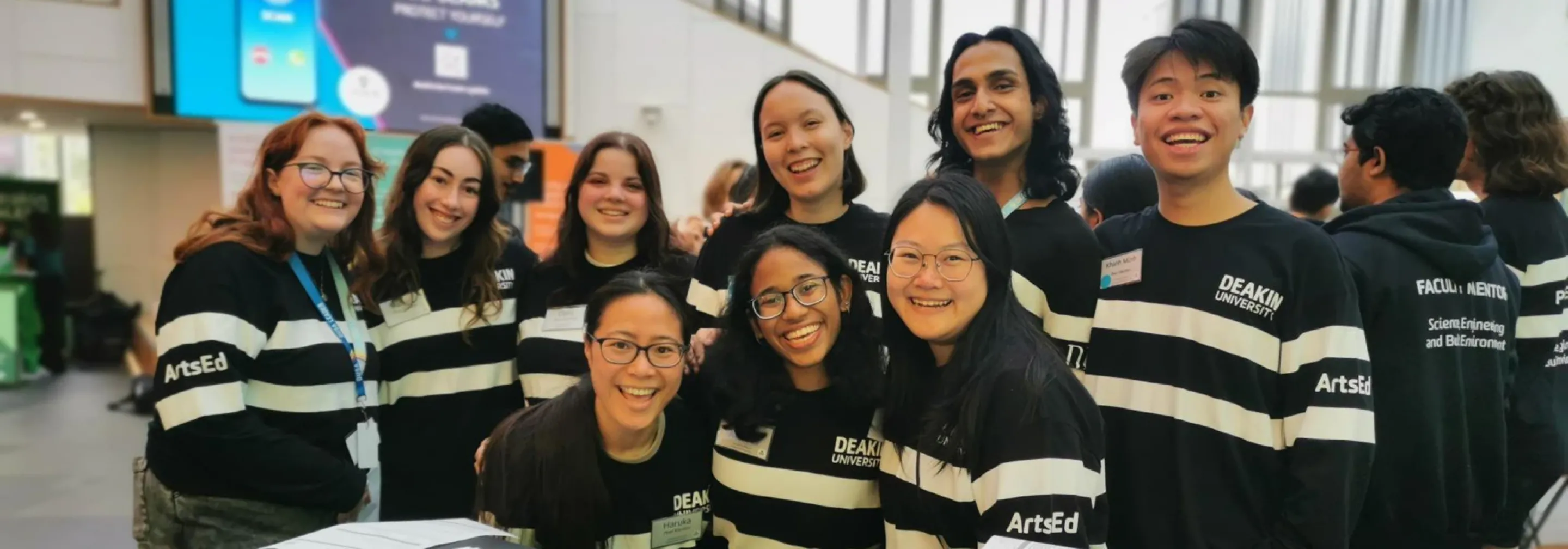 A group photo of nine peer mentors, gathered together smiling at the camera. They are wearing matching branded jumpers, with the word ArtsEd on the sleeve.