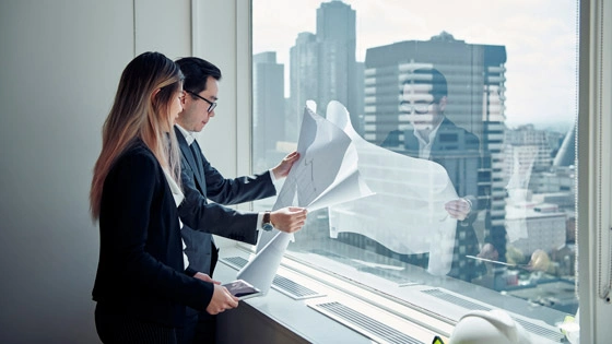 Two students look at building plans in front of a window