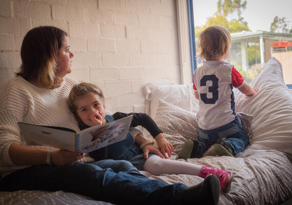 A mother sits on a bed holding a book with two young children 