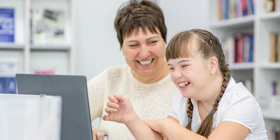 A woman observes a young child with a disability on a laptop. They are both smiling.