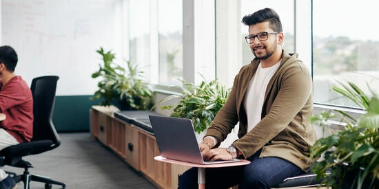Student smiles while working at a laptop next to a meeting.