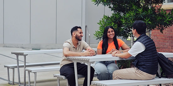 Three students sitting outside at a metal table, talking and laughing