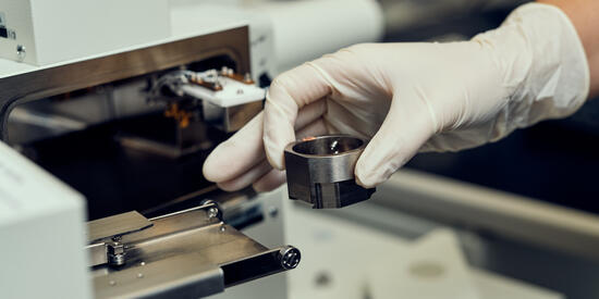 Researcher's gloved hands holding a metal object in front of a machine in the Institute for Frontier Materials