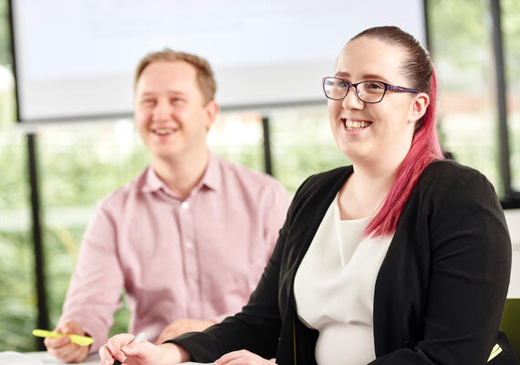 Two people holding pens and smiling inside of a classroom.