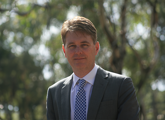 A close up of Craig Olsson, wearing a suit smiling at the camera against a blue background.