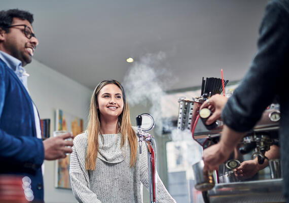 A cafe scene with to people waiting for a coffee standing next to a coffee machine