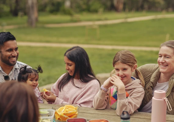 Several families sit together outside on a park bench enjoying a picnic.