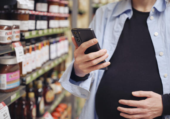 A pregnant person holding a shopping basket and using their phone.