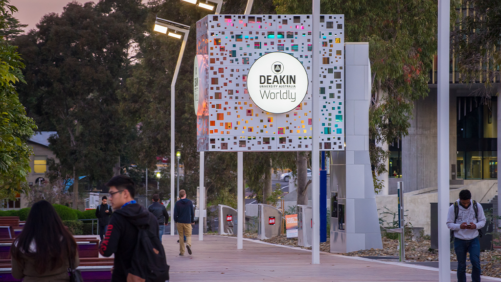 Students walk through Deakin University's Burwood Campus at dusk, passing an illuminated Deakin sign along a pedestrian pathway lined with trees, seating and campus buildings.