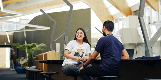 Two Deakin students laughing and talking while sitting on stools at Student Central at the Melbourne Burwood Campus.