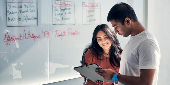 Two students in front of a whiteboard looking at documents.