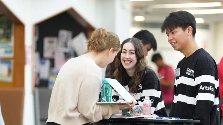 Two people stand smiling at a third person with her back to the camera. They are wearing jumpers with the word ArtsEd on the sleeve.