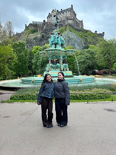 Ellie and a friend pose smiling in front of a teal fountain.