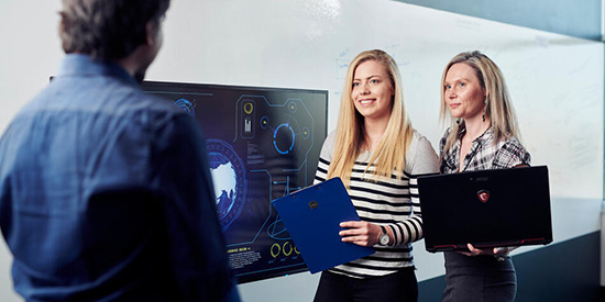 Two Deakin computer science student stand holding a laptop and clipboard.