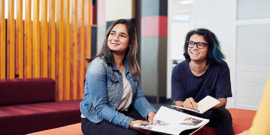 Two Deakin Burwood Campus students sit on a sofa together, looking at a book and smiling.