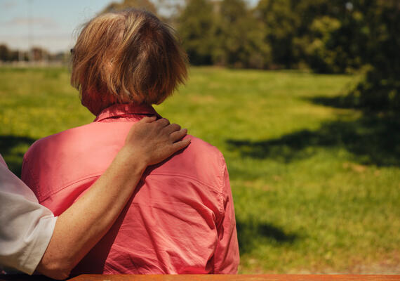 A person sits on a park bench and a family member puts their arm around their shoulders.