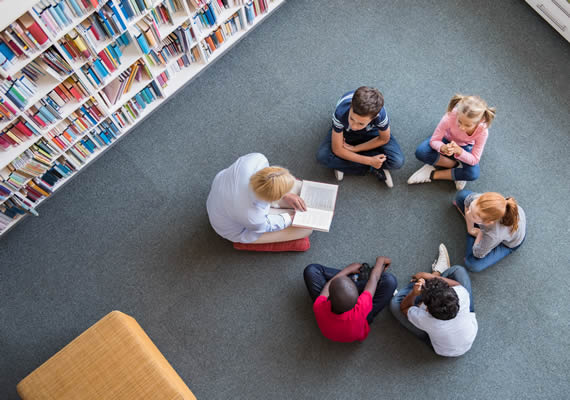 Children sitting in circle in a library with a teacher. Teacher is reading a book.