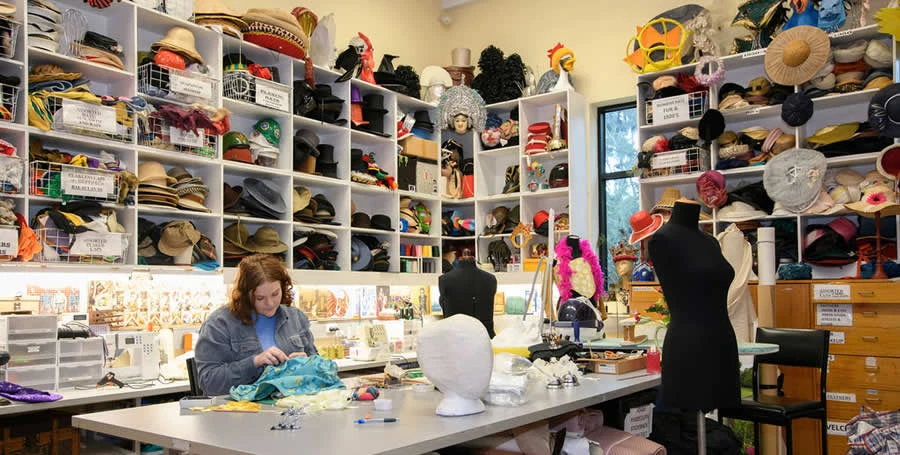 The wardrobe department, showing a person sitting at table and tailoring a costume, and room filled with colourful hats and costumes