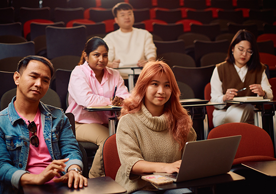 A group of five students with study materials seated across three rows in a lecture theatre, looking towards the front of the room.