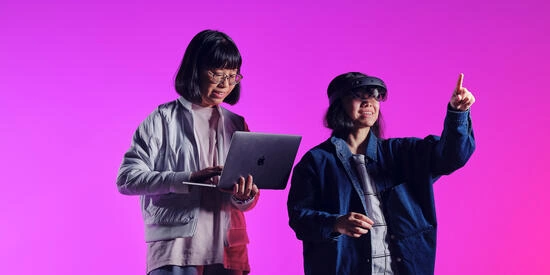 Two information technology course students. One is holding a laptop and the other is wearing a VR headset. The background is bright pink.